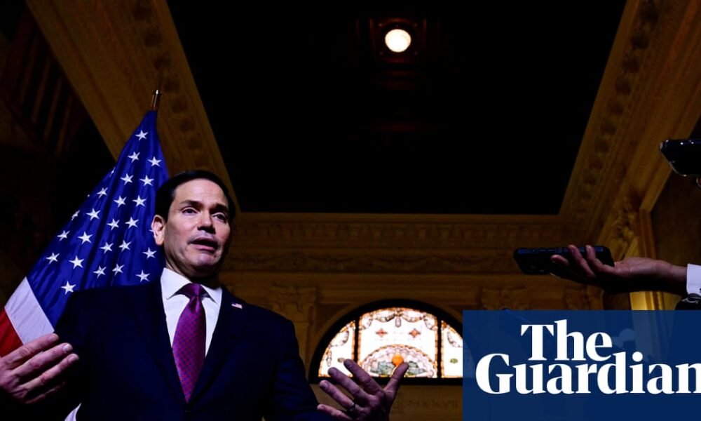 Marco Rubio — Un hombre en traje habla en un evento, con la bandera de Estados Unidos visible detrás de él mientras asistentes toman fotografías. Foto de: The Guardian