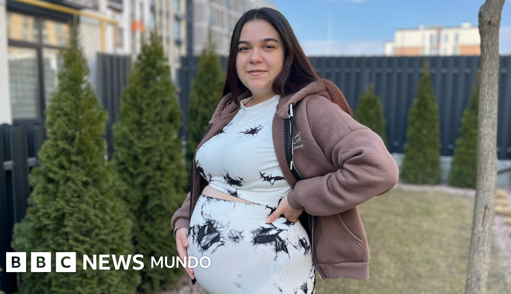 Ucrania debate ley — Una mujer embarazada está de pie en un jardín con la mano sobre el vientre. Tiene el pelo largo, liso y oscuro, y lleva un conjunto de blusa y pantalón blancos con detalles negros. También lleva una sudadera con capucha de color marrón claro.
