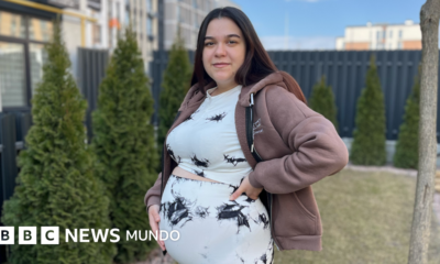 Ucrania debate ley — Una mujer embarazada está de pie en un jardín con la mano sobre el vientre. Tiene el pelo largo, liso y oscuro, y lleva un conjunto de blusa y pantalón blancos con detalles negros. También lleva una sudadera con capucha de color marrón claro.