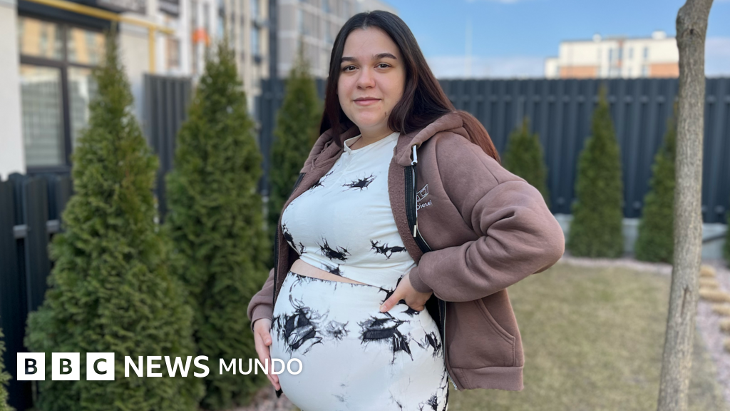Ucrania debate ley — Una mujer embarazada está de pie en un jardín con la mano sobre el vientre. Tiene el pelo largo, liso y oscuro, y lleva un conjunto de blusa y pantalón blancos con detalles negros. También lleva una sudadera con capucha de color marrón claro.