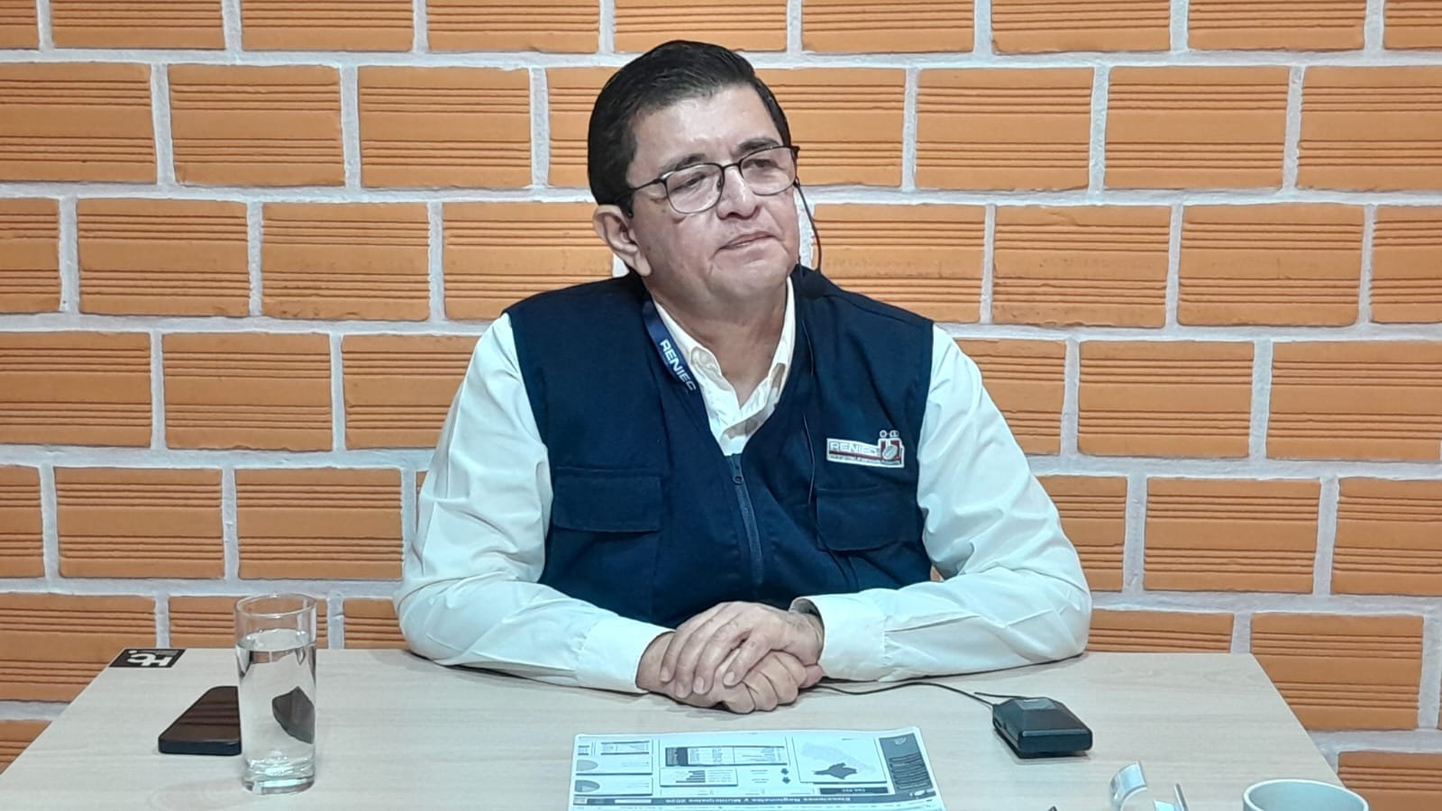 padrón electoral — Un hombre con gafas y chaleco azul sentado ante una mesa con documentos y un vaso de agua frente a una pared de ladrillos. Foto de: El Búho