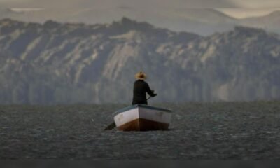 documental uyariy — Un hombre con sombrero y ropa oscura permanece de pie en una pequeña barca sobre aguas tranquilas con montañas al fondo. Foto de: La República