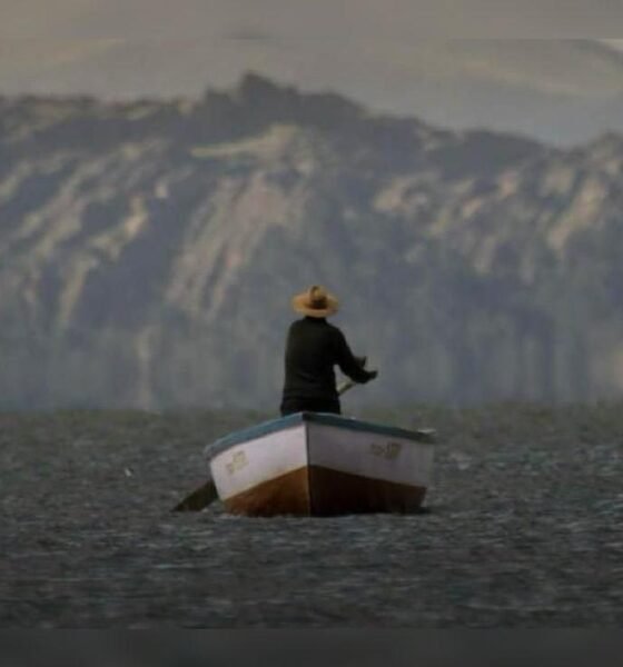 documental uyariy — Un hombre con sombrero y ropa oscura permanece de pie en una pequeña barca sobre aguas tranquilas con montañas al fondo. Foto de: La República