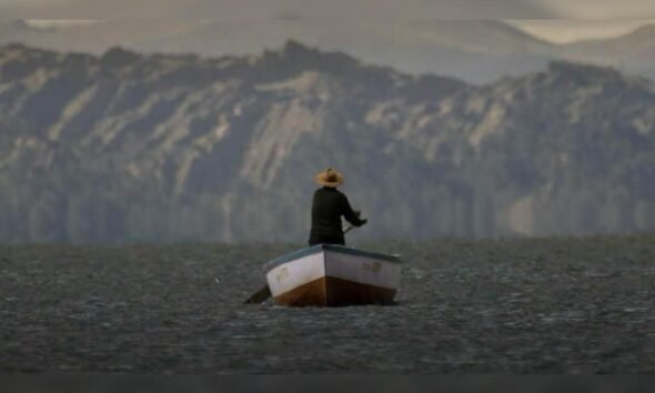 documental uyariy — Un hombre con sombrero y ropa oscura permanece de pie en una pequeña barca sobre aguas tranquilas con montañas al fondo. Foto de: La República