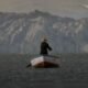 documental uyariy — Un hombre con sombrero y ropa oscura permanece de pie en una pequeña barca sobre aguas tranquilas con montañas al fondo. Foto de: La República