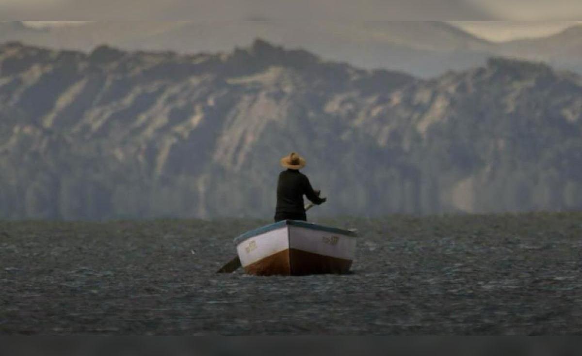 documental uyariy — Un hombre con sombrero y ropa oscura permanece de pie en una pequeña barca sobre aguas tranquilas con montañas al fondo. Foto de: La República