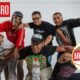 Nicky Jam — Un grupo de hombres posan sonrientes frente a un fondo blanco con logotipos y un anuncio publicitario. Foto de: El Búho