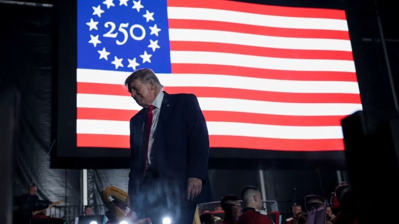 Donald Trump aparece frente a una bandera conmemorativa del 250 aniversario de Estados Unidos en medio de las quejas por la organización del evento. Foto de: CNN en Español