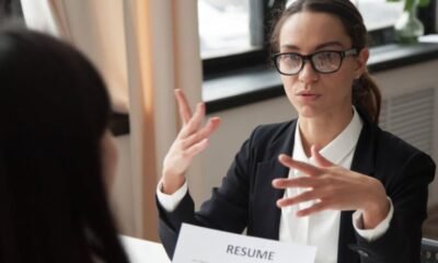 Empresas restringen aumentos — Una mujer con gafas y traje oscuro gesticula mientras habla con otra persona frente a un documento que dice 'RESUME'. Foto de: Gestión