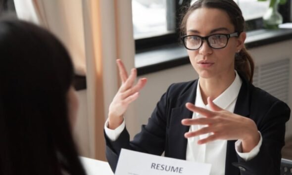 Empresas restringen aumentos — Una mujer con gafas y traje oscuro gesticula mientras habla con otra persona frente a un documento que dice 'RESUME'. Foto de: Gestión