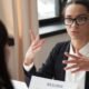 Empresas restringen aumentos — Una mujer con gafas y traje oscuro gesticula mientras habla con otra persona frente a un documento que dice 'RESUME'. Foto de: Gestión