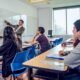 requisitos establecidos — Un profesor señala una gráfica en una pizarra mientras tres estudiantes escuchan en un salón de clases. Foto de: El Peruano