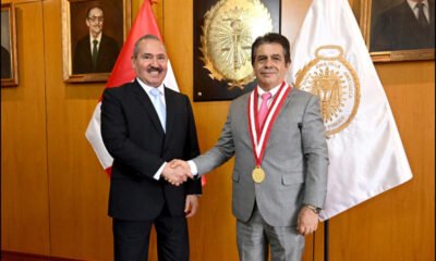 crimen organizado — Dos hombres con traje, uno con una medalla roja, se dan la mano frente a banderas y retratos. Foto de: El Peruano